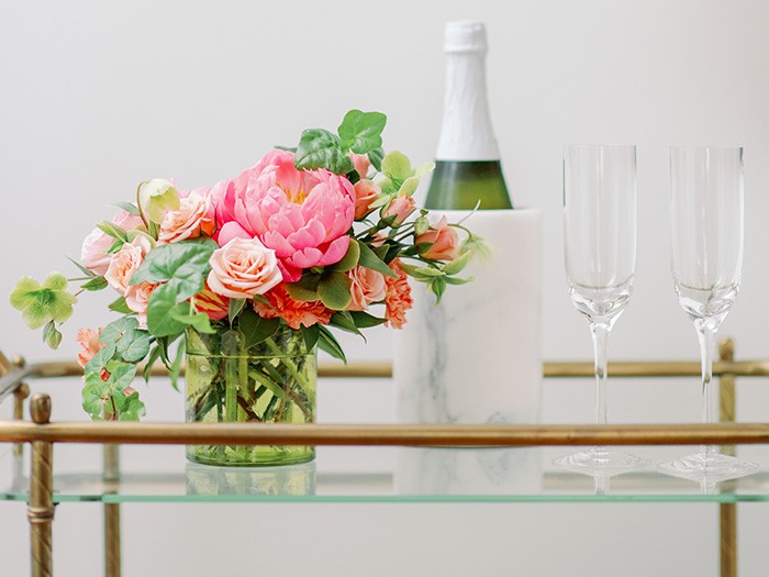 Gold entertainment cart with pink flowers in round glasss vase, crystal wine glasses and white marble holder with champagne bottle. 